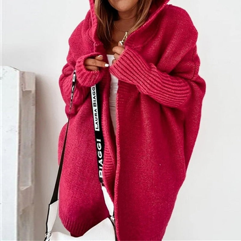 Femme portant un long cardigan fuchsia en maille épaisse, avec capuche et manches côtelées, enveloppant sa tenue blanche dans un décor lumineux et minimaliste.