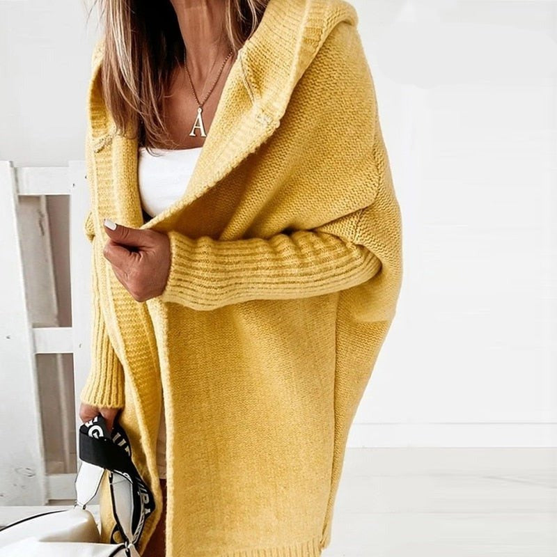 Femme portant un long cardigan jaune en maille douce, doté d’une capuche et de manches côtelées, superposé à une tenue blanche dans un décor lumineux et minimaliste.