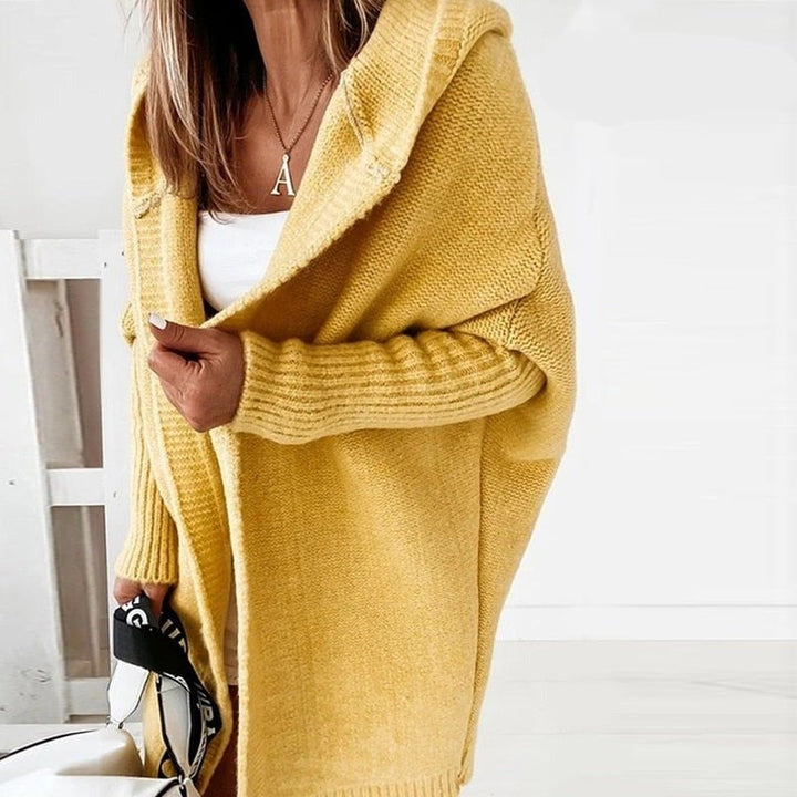 Femme portant un long cardigan jaune en maille douce, doté d’une capuche et de manches côtelées, superposé à une tenue blanche dans un décor lumineux et minimaliste.