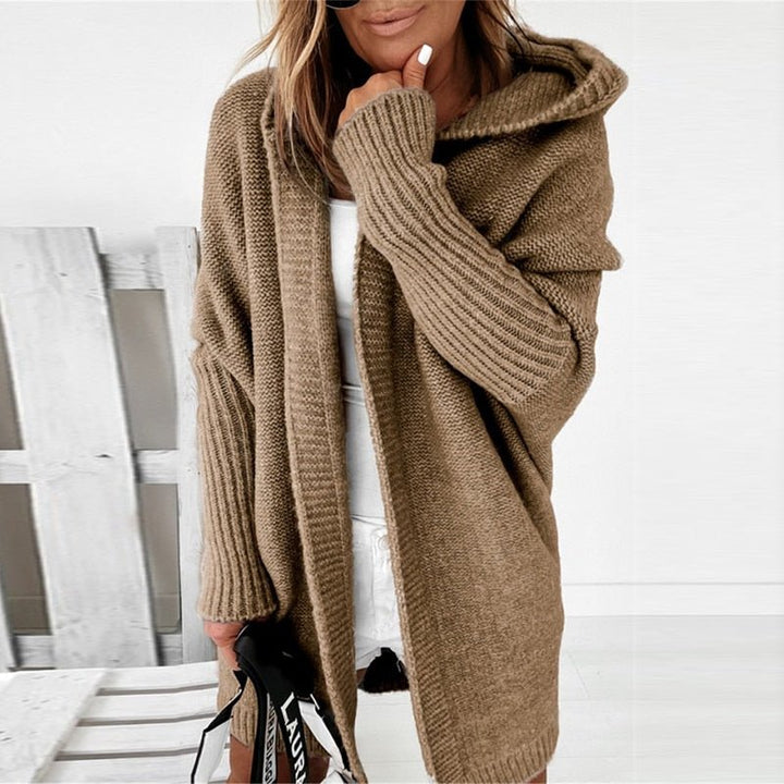 Femme portant un long cardigan beige en maille épaisse, avec capuche et manches côtelées, associé à une tenue blanche dans un décor clair et minimaliste.