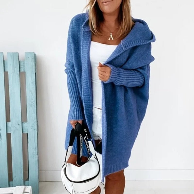 Femme portant un long cardigan bleu en maille épaisse, doté d’une capuche et de manches côtelées, superposé à une tenue blanche dans un décor lumineux et minimaliste.