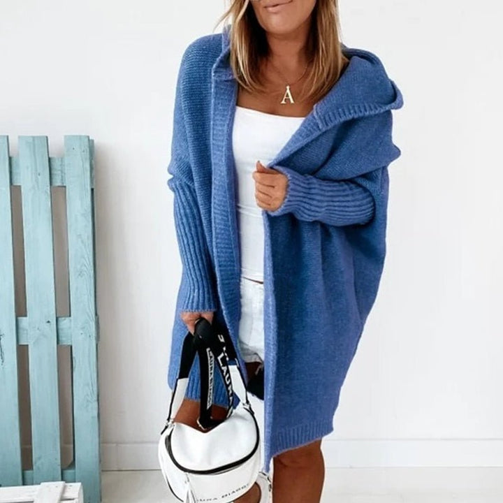 Femme portant un long cardigan bleu en maille épaisse, doté d’une capuche et de manches côtelées, superposé à une tenue blanche dans un décor lumineux et minimaliste.