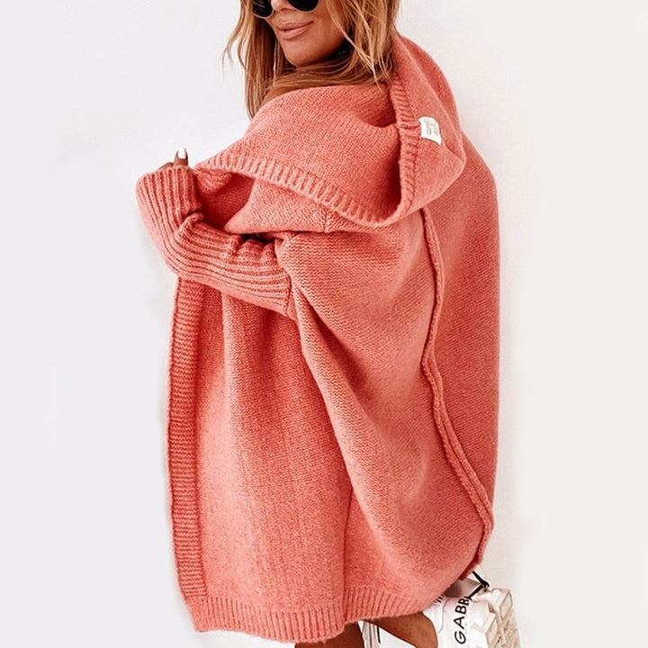 Femme portant un long cardigan en maille épaisse de couleur corail, avec capuche et manches côtelées, présenté dans un décor clair et minimaliste.