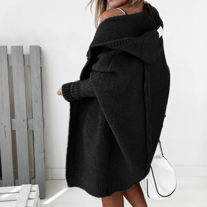 Femme portant un long cardigan noir en maille épaisse, avec capuche et manches côtelées, dévoilant une épaule, dans un décor lumineux et minimaliste.