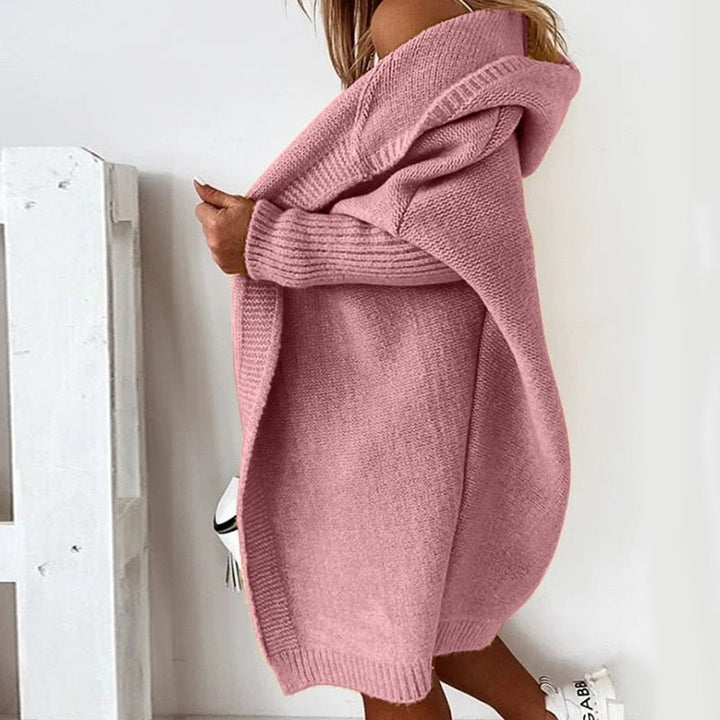 Femme portant un long cardigan rose en maille épaisse, avec capuche et manches côtelées, laissant légèrement apparaître une épaule, dans un décor clair et minimaliste.
