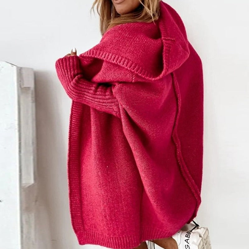 Femme portant un long cardigan fuchsia en maille épaisse, avec capuche et manches côtelées, dévoilant légèrement une épaule dans un décor minimaliste et lumineux.