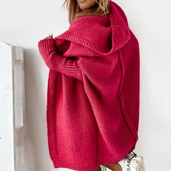 Femme portant un long cardigan fuchsia en maille épaisse, avec capuche et manches côtelées, dévoilant légèrement une épaule dans un décor minimaliste et lumineux.