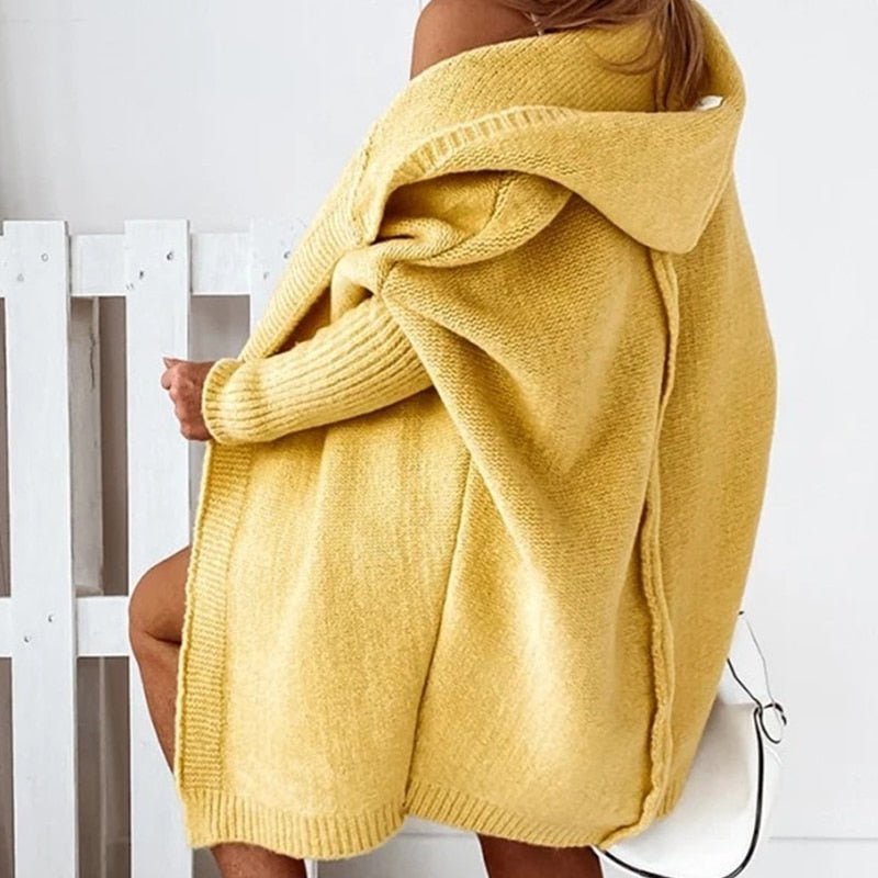 Femme portant un long cardigan jaune en maille épaisse, avec capuche et manches côtelées, dévoilant légèrement une épaule, dans un décor clair et minimaliste.