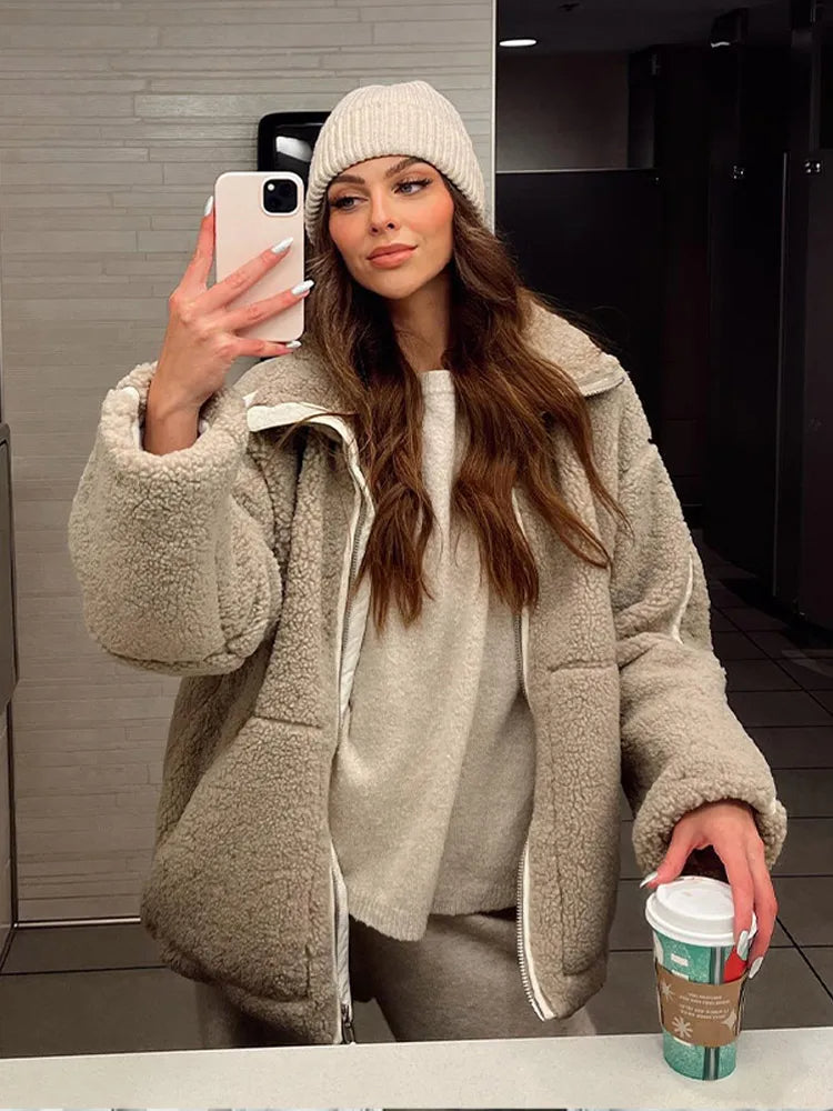 Femme portant une veste teddy beige avec une fermeture éclair, un pull assorti et un pantalon de survêtement. Elle tient un gobelet de café et porte un bonnet en tricot. L’image montre une allure décontractée et cosy, parfaite pour l’hiver, dans un cadre intérieur moderne.