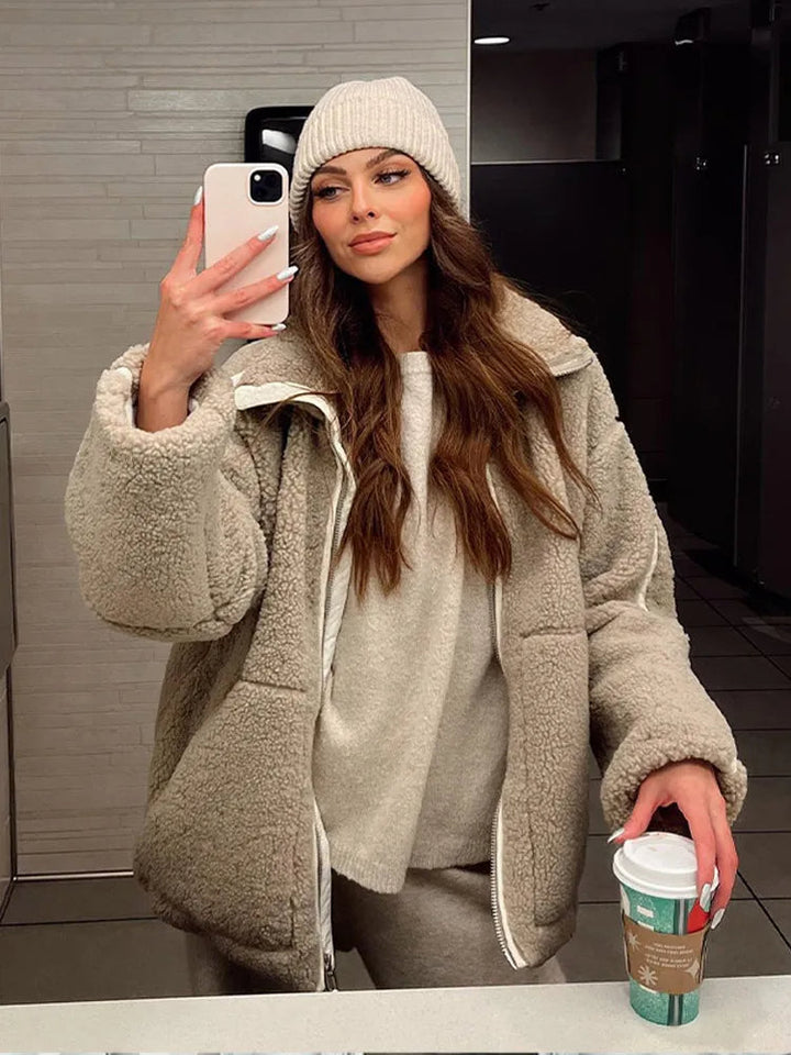 Femme portant une veste teddy beige avec une fermeture éclair, un pull assorti et un pantalon de survêtement. Elle tient un gobelet de café et porte un bonnet en tricot. L’image montre une allure décontractée et cosy, parfaite pour l’hiver, dans un cadre intérieur moderne.
