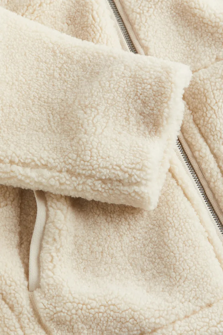 Un gros plan sur la manche et la fermeture éclair d'une veste en peluche blanche révèle la texture douce et moelleuse du tissu. Le détail de la fermeture éclair et la bordure en tissu contrastant au col soulignent le confort et la qualité de la matière.