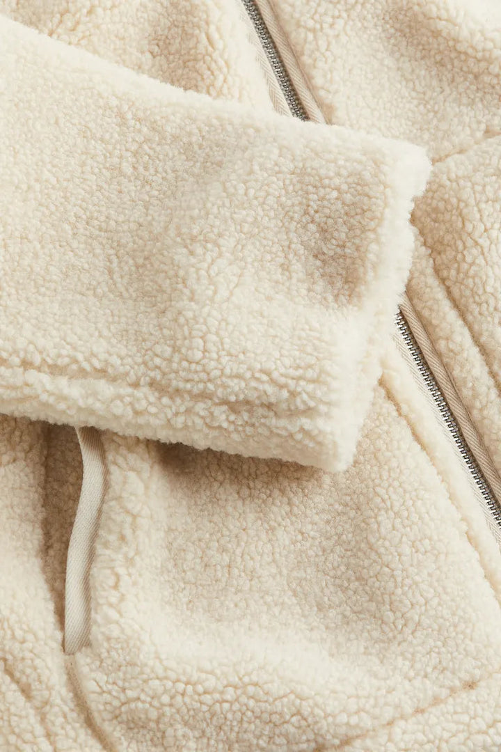 Un gros plan sur la manche et la fermeture éclair d'une veste en peluche blanche révèle la texture douce et moelleuse du tissu. Le détail de la fermeture éclair et la bordure en tissu contrastant au col soulignent le confort et la qualité de la matière.