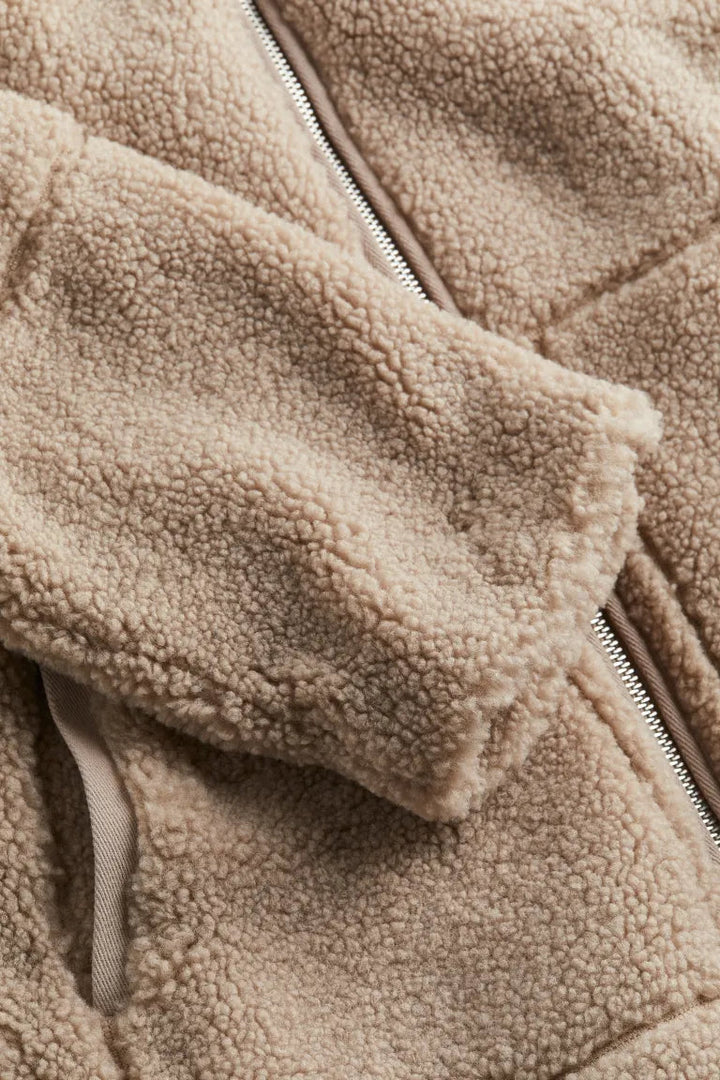 Un gros plan sur la manche et la fermeture éclair d'une veste en peluche marron met en valeur la texture douce du tissu. L'image révèle les détails de la matière et la finition de la fermeture éclair, illustrant le confort et la qualité de la confection.