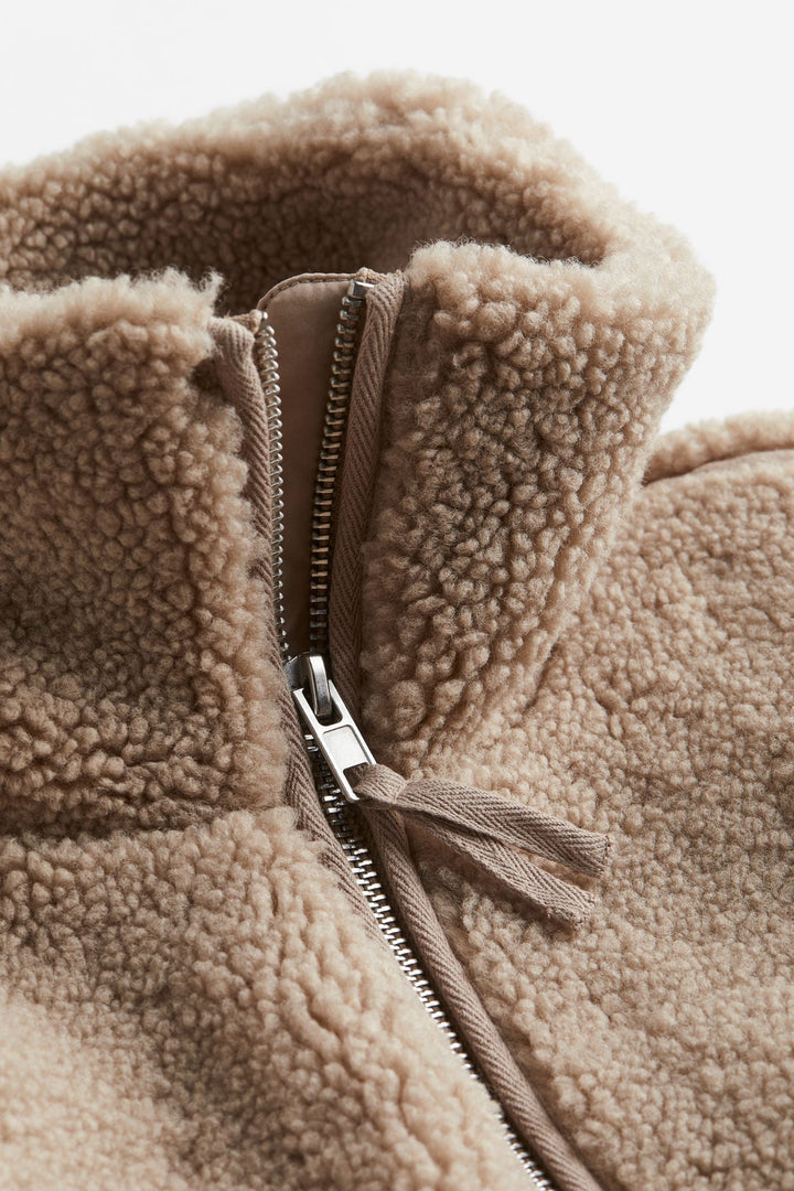 Un gros plan sur la fermeture éclair d'une veste teddy marron met en valeur le détail de la fermeture argentée et la douceur du tissu pelucheux. Le col montant et les finitions soignées de la veste sont également visibles, soulignant son confort et sa qualité.